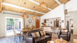 A spacious living area with a leather sofa, dining table, and chairs. There are pendant lights, a kitchen in the background, and a large window. - Villa La Roche Rouge (Pougnadoresse)