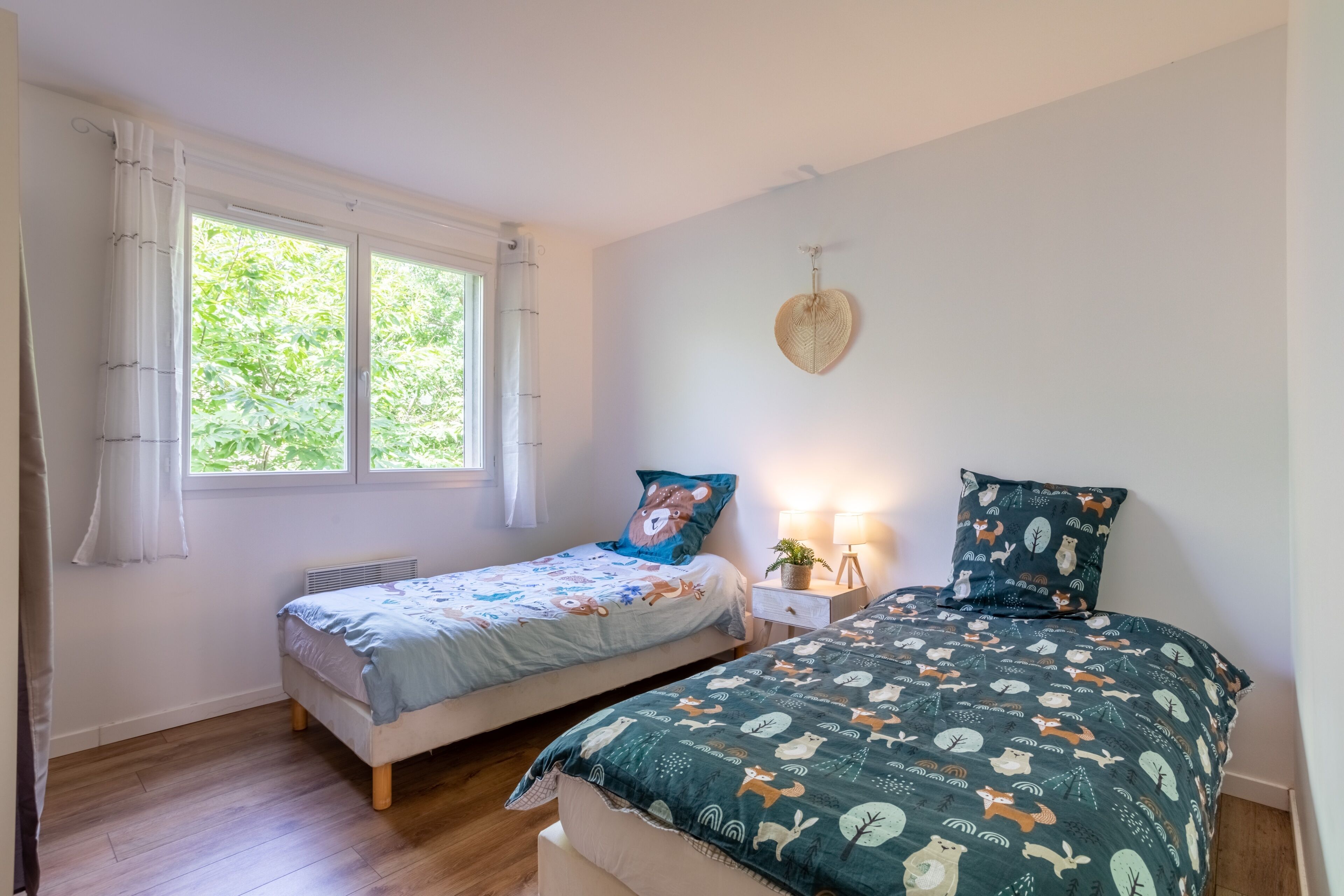 A bedroom with two beds, a window with curtains, a bedside table with a lamp, and a wall-mounted decorative item.