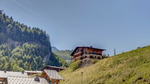 A mountain lodge with multiple balconies, surrounded by forested hills and a clear sky. - Le Col des Aravis - Two Bedroom Apartment, Sleeps 5 (La Giettaz)