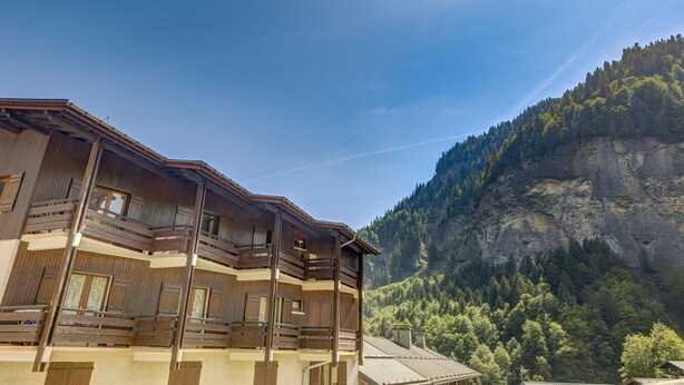 A wooden chalet with multiple balconies, set against a backdrop of a forested mountain. - Le Col des Aravis - Two Bedroom Apartment, Sleeps 5 (La Giettaz)