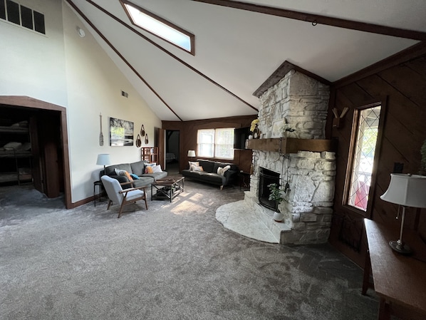A spacious living room with a stone fireplace, a lofted ceiling, and a large window providing natural light.