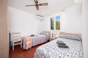 Ein Schlafzimmer mit zwei Betten, einem Deckenventilator, einer Klimaanlage, einem Fenster mit Vorhängen und einem kleinen Tisch mit einer Flasche und einer Lampe.