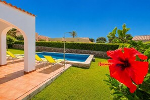 A swimming pool with a white pergola and yellow lounge chairs, surrounded by well-manicured grass and a red hibiscus flower. - Villa Gelabert Ametller (Ciutadella de Menorca)