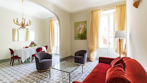 A living room with a red sofa, two armchairs, a glass coffee table, and a chandelier. There is a dining area with a table and chairs, and a painting of flowers on the wall. - Capo 10 Liberty C3 - Two Bedroom Apartment, Sleeps 4 (Syracuse)