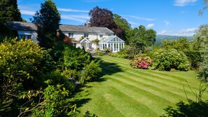 A large house with a well-maintained lawn and garden, surrounded by trees and shrubs. - Orchard Cottage (Ambleside)