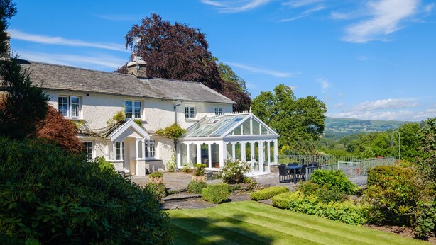 A large, white, two-story house with a conservatory, surrounded by well-maintained gardens and a clear view of the countryside. - Orchard Cottage (Ambleside)