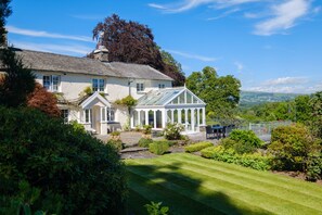 A large, white, two-story house with a conservatory, surrounded by well-maintained gardens and a clear view of the countryside. - Orchard Cottage (Ambleside)
