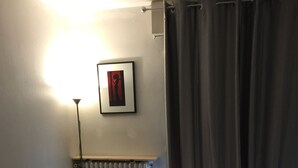 A bedroom with a bed, a red pillow, a radiator, a picture on the wall, and a curtain. - Logement Clair ET Spacieux (Paris)