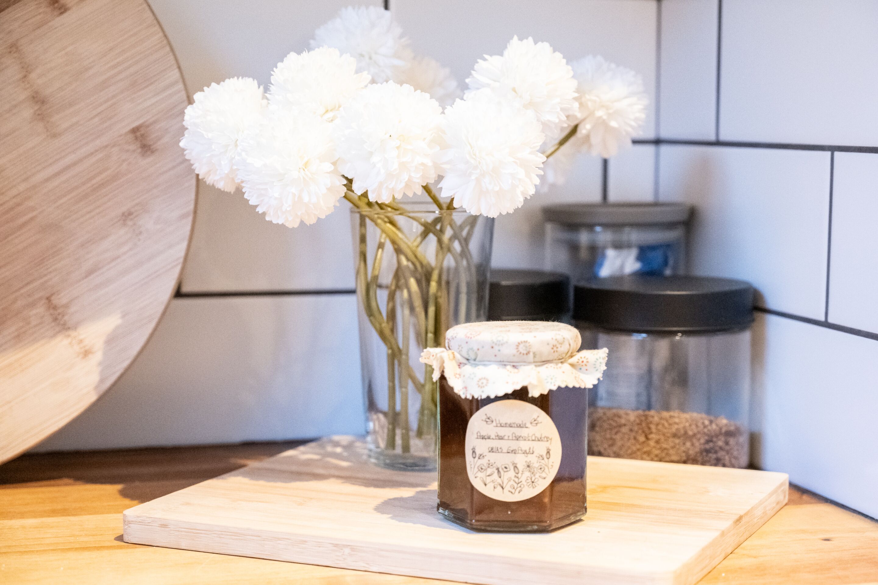 En køkkenbordplade med et glas hjemmelavet marmelade og en vase med hvide blomster.