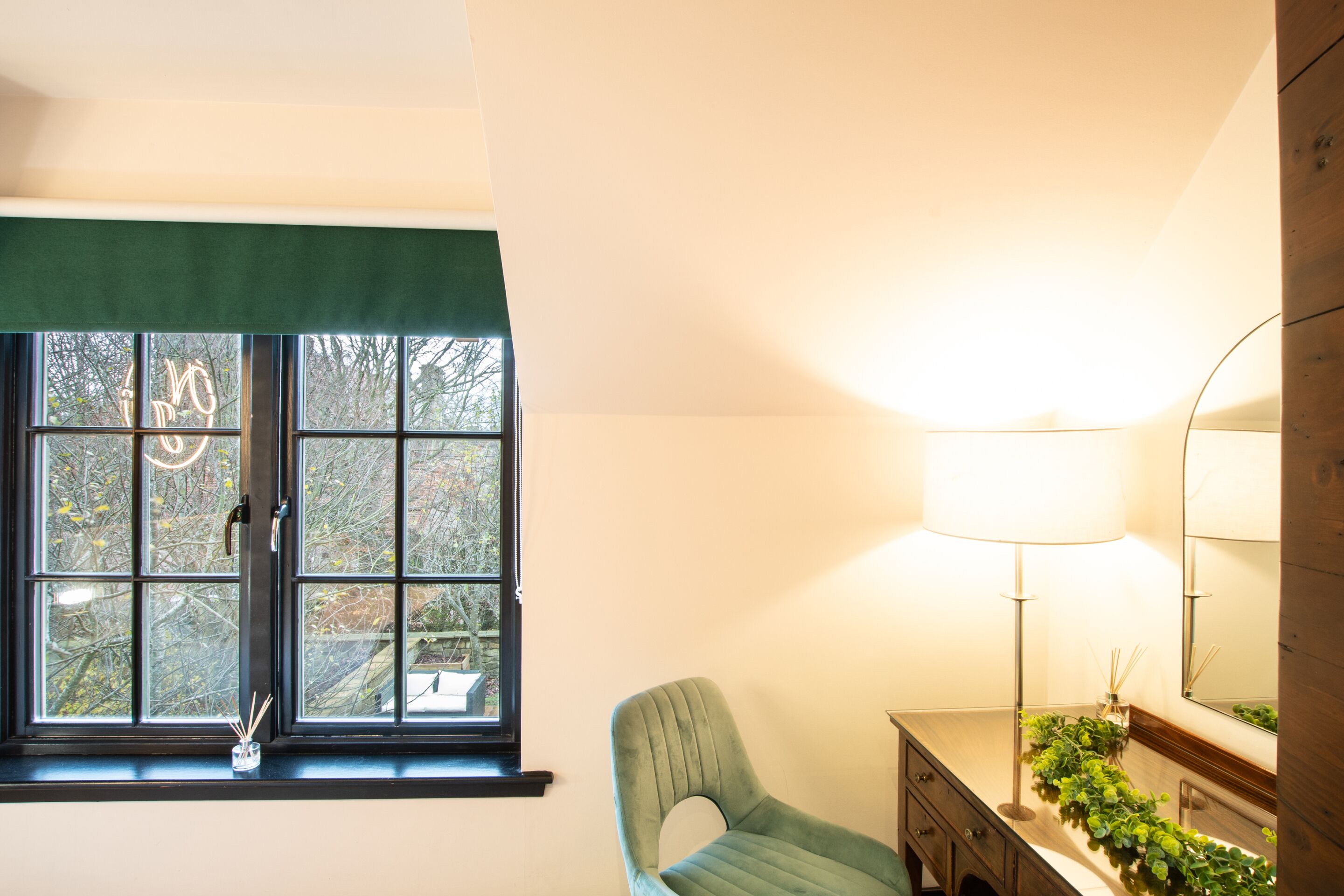 Une pièce avec une fenêtre, un fauteuil vert, une table de chevet, une lampe et un miroir.