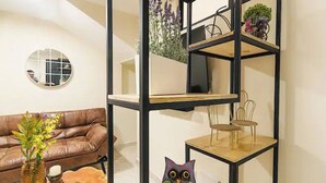 A modern living room with a brown leather sofa, a round mirror, a wooden coffee table with a vase and candle, and a black metal shelving unit with plants and a television. - Cozy corner in El Poblado! (Medellín)