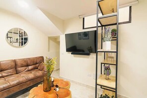 A living room with a brown leather couch, a round coffee table, a TV mounted on the wall, and a wooden bookshelf with decorative items. - A cozy little spot in El Poblado! (Medellín)