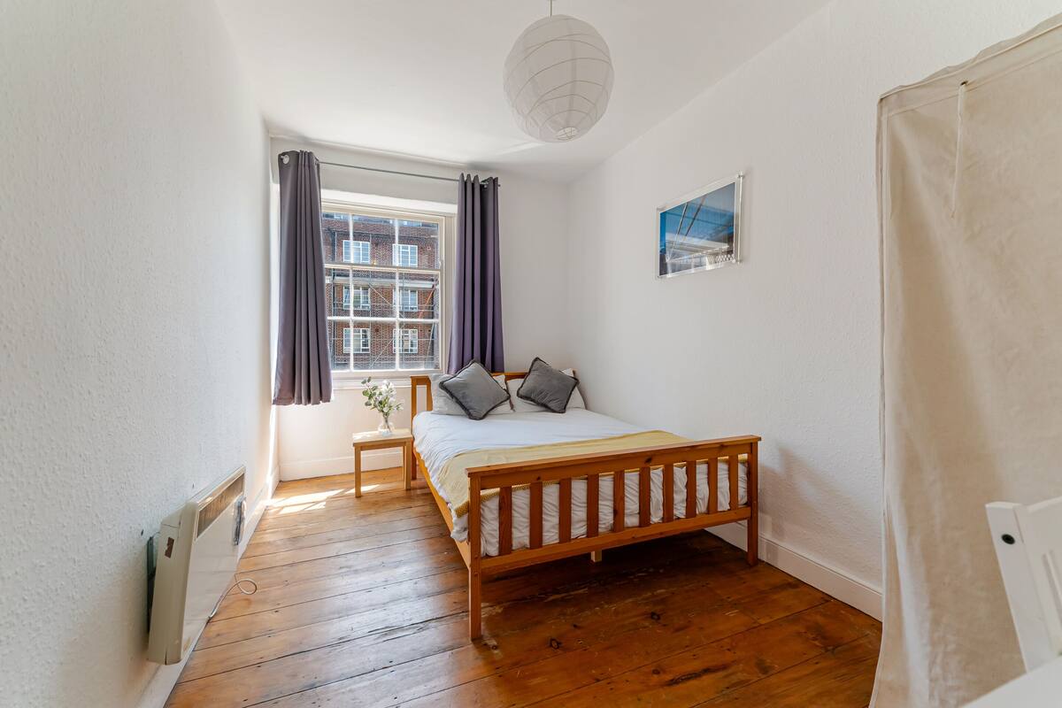 Een slaapkamer met een houten bed, een raam met gordijnen, een radiator en een foto aan de muur.