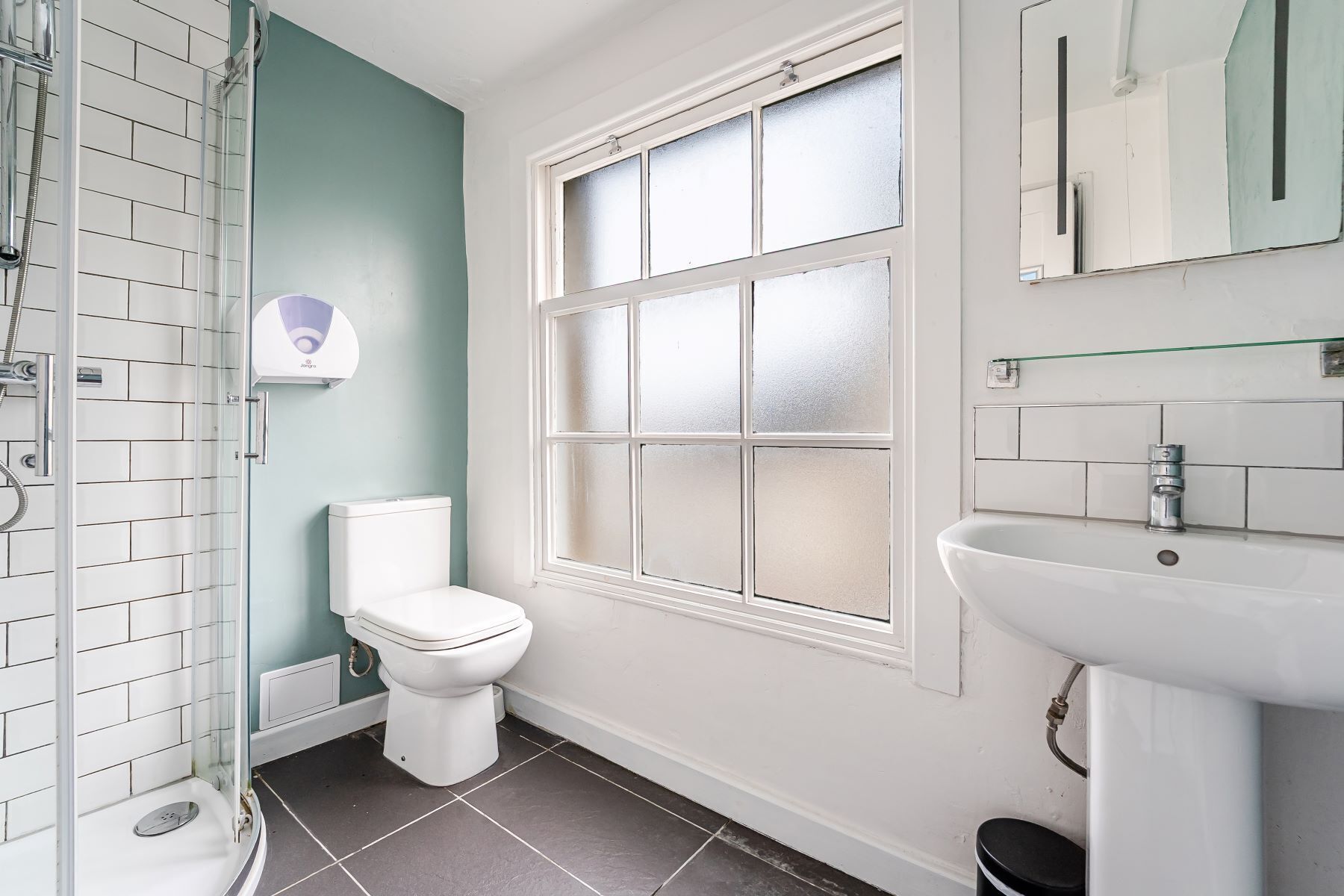 A bathroom with a white toilet, a glass shower enclosure, a window with frosted glass, and a white sink with a mirror above it.