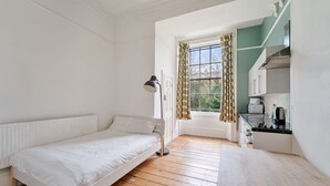 Een kleine, schone kamer met een bed, bureau, lamp, raam met gordijnen en een kitchenette.
