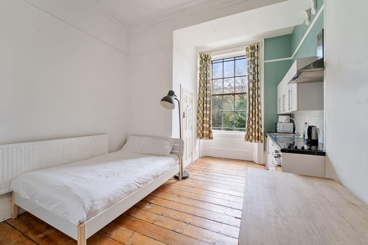 Een kleine, schone kamer met een bed, bureau, lamp, raam met gordijnen en een kitchenette.