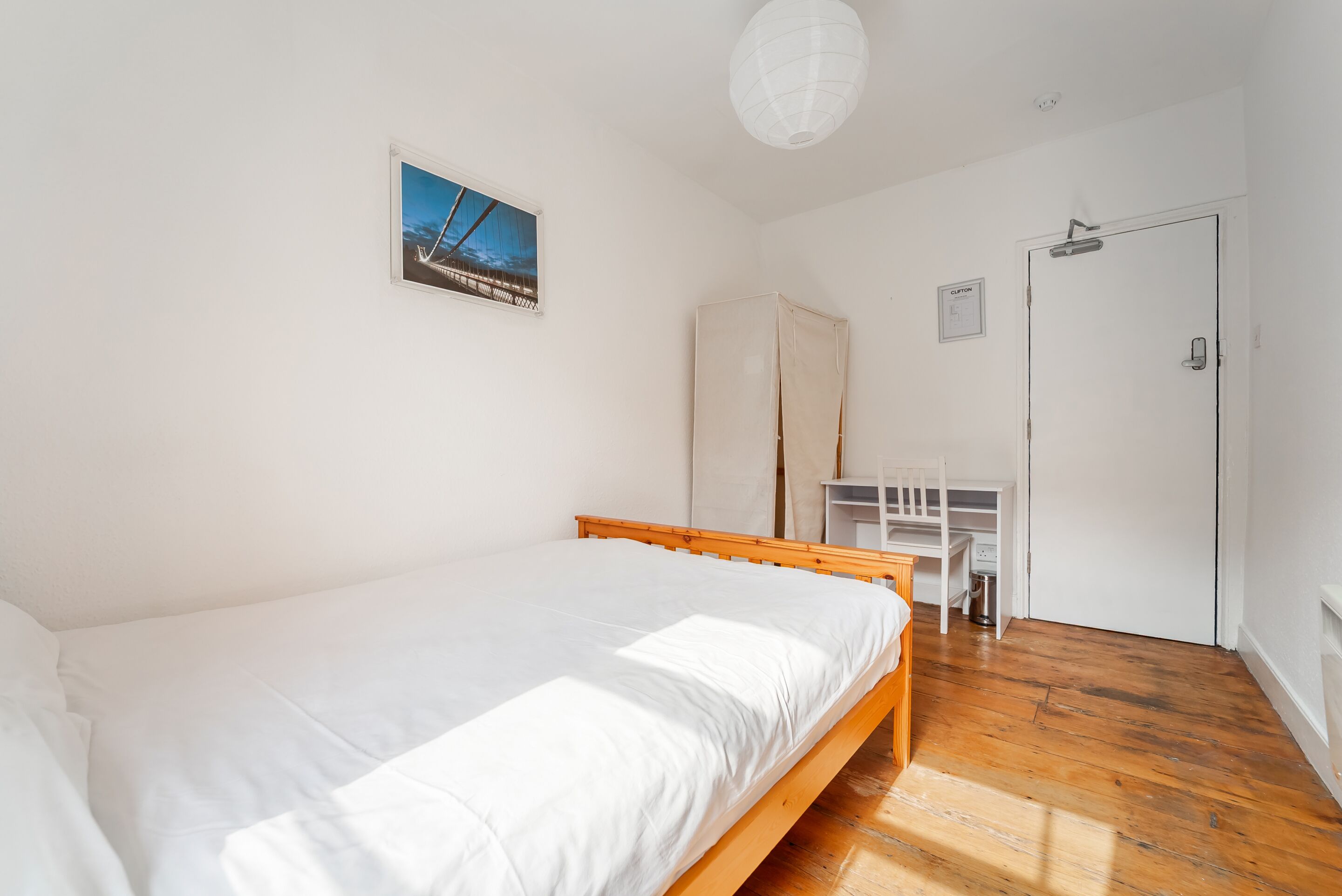 A bedroom with a wooden bed, a desk, a chair, a wardrobe, and a door.