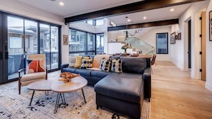 A modern living room with a sectional sofa, a round coffee table, and a large window offering a view of the city. - The Ridgeline | Ski-In 4BR Mountain Contemporary (Park City)