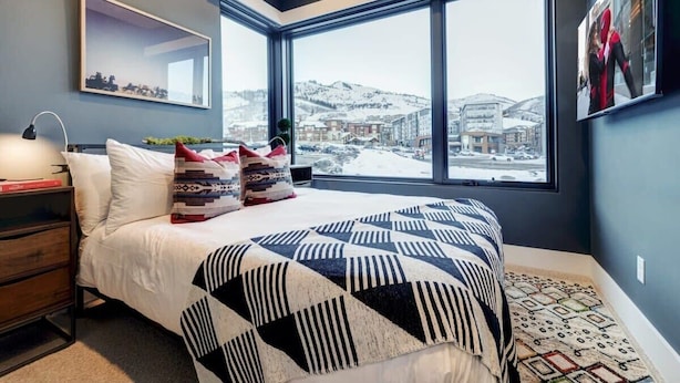 A bedroom with a large bed, a nightstand, a lamp, a window with a view of a snowy landscape, and a framed picture on the wall. - The Ridgeline | Ski-In 4BR Mountain Contemporary (Park City)