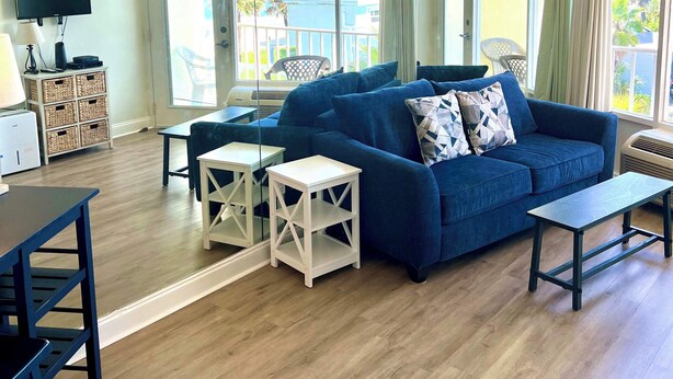 A spacious living room with a blue sofa, white side tables, and a view of the ocean through large windows. - Waters Edge  - Ocean View at Symphony Beach Club! (Ormond Beach)