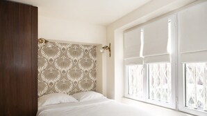 Un dormitorio con cama, cabecera con diseño estampado, ventana con persianas y lámpara fijada a la pared.