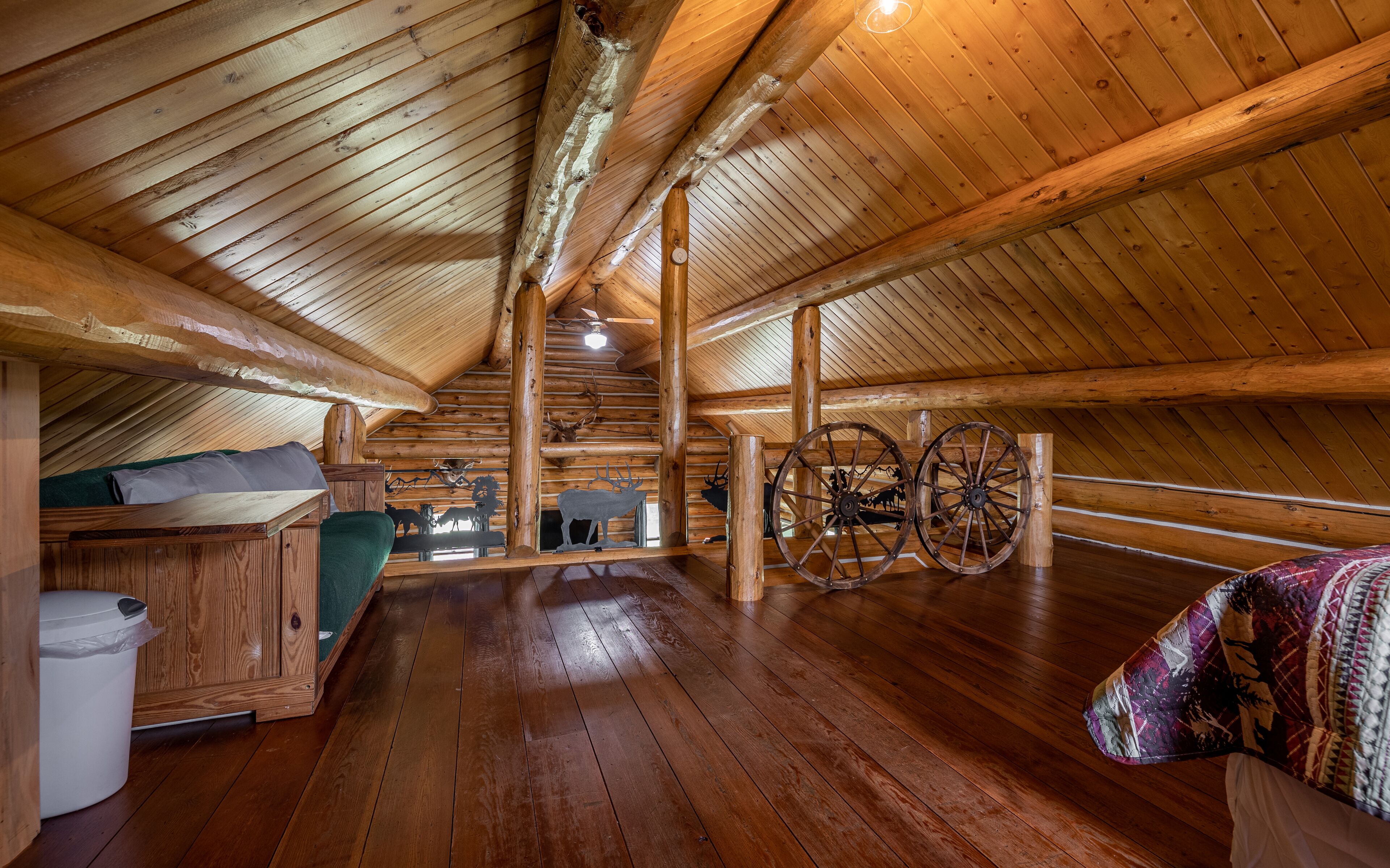 Interior de una cabaña de troncos con piso de madera, una cama, un banco, un ventilador de techo y una rueda decorativa.