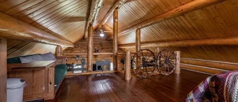 Interior de una cabaña de troncos con piso de madera, una cama, un banco, un ventilador de techo y una rueda decorativa.