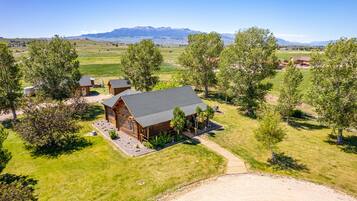 A log cabin with a metal roof, surrounded by a fenced yard and trees, with a mountain range in the background.