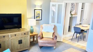 A living room with a television, a brown leather armchair, a blue sofa, a wooden coffee table, and a dining area with a table and chairs. - Inviting Townhouse in Bedlington (Bedlington)