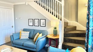 A living room with a blue sofa, a wooden coffee table, a small side table with a lamp, and a staircase with a white railing. - Inviting Townhouse in Bedlington (Bedlington)