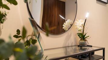 Un baño con tocador de vidrio, un espejo redondo, una orquídea en maceta y una puerta de madera al fondo.