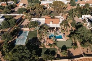 An aerial view of a residential area with a house featuring a swimming pool, tennis court, and landscaped garden.