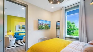 A hotel room with a yellow bedspread, a large window with a view of greenery, a flat-screen TV on the wall, and a bright yellow wall in the adjacent room.