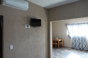 A room with beige walls, a flat-screen TV mounted on the wall, a wooden table, and a patterned curtain. - Bungalows Victoria (La Manzanilla)