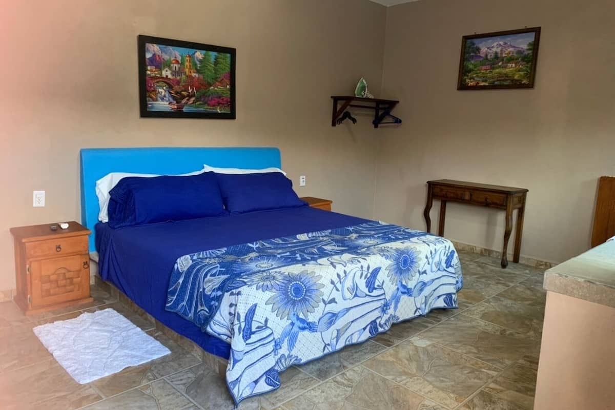 A bedroom with a blue bedspread, a wooden nightstand, a small table, and two wall-mounted shelves.