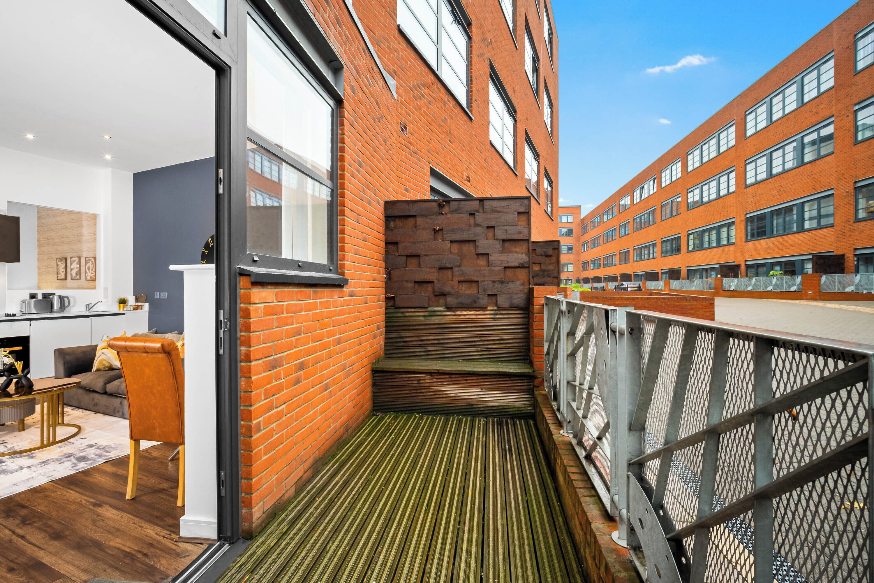 Un appartement moderne comprenant une cuisine, un espace de vie avec un canapé et une table basse, ainsi qu’un balcon doté d’un plancher en bois et d’une rambarde en métal.