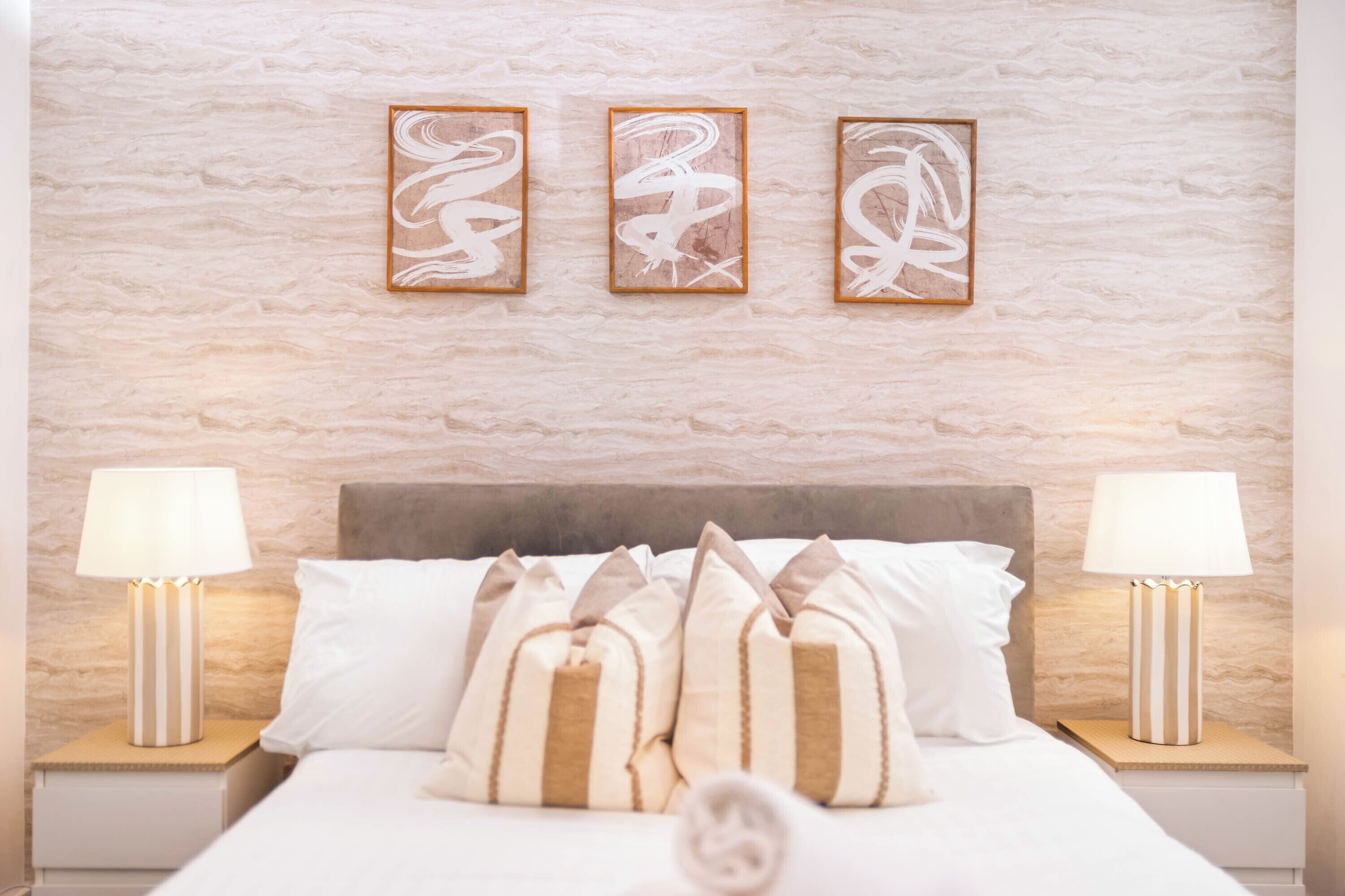 A neatly arranged hotel room with a bed, pillows, two bedside lamps, and three framed artworks on the wall.