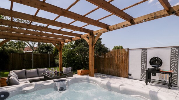 Ein Whirlpool unter einer Hollaube in einem Hinterhof mit Holzzaun, Grill und Gartenmöbeln.