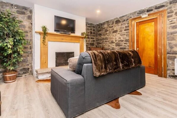 A cozy living room with a grey sofa, a fireplace, a television, and a stone wall. - Modern Meets Rustic - 2 Bdrm Old Quebec (Québec City)
