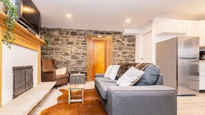 A cozy living room with a stone wall, a fireplace, a grey sofa, and a kitchen area with white cabinets. - Modern Meets Rustic - 2 Bdrm Old Quebec (Québec City)