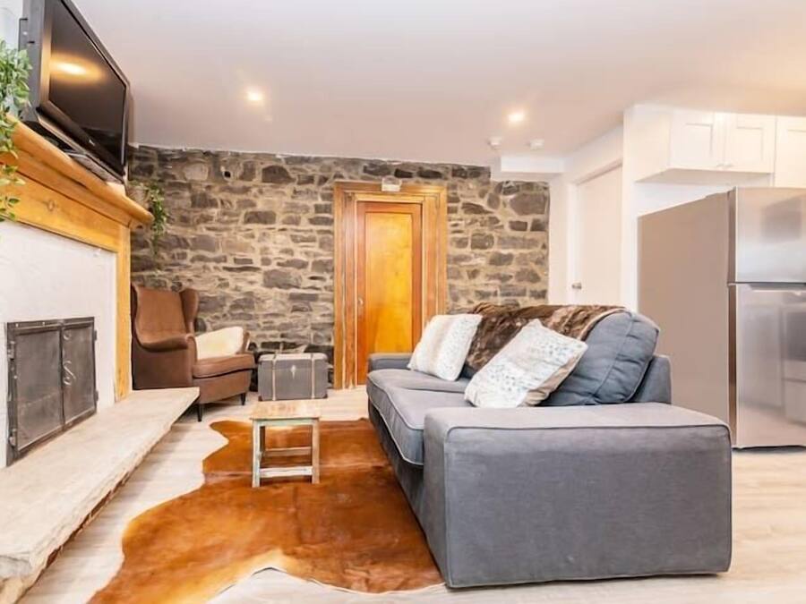 A cozy living room with a stone wall, a fireplace, a grey sofa, and a kitchen area with white cabinets.