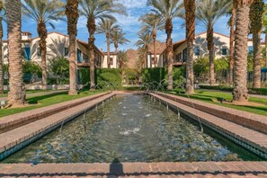 A rectangular pool with clear water, surrounded by palm trees and buildings with red-tiled roofs. - The Luxe Loft A | 1BR Legacy Villa (La Quinta)
