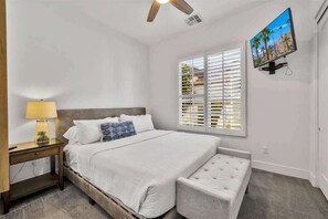 A bedroom with a bed, a nightstand, a lamp, a window with blinds, a ceiling fan, and a TV mounted on the wall.