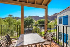 A covered outdoor seating area with a table and chairs, overlooking a mountainous landscape with greenery and a clear sky.