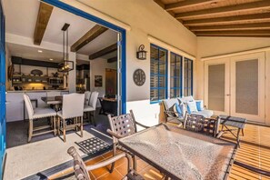 A spacious outdoor dining area with a table and chairs, a kitchen visible through an open door, and a seating area with cushions. - The Luxe Loft A | 1BR Legacy Villa (La Quinta)
