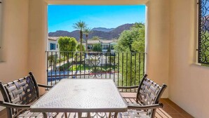 Un balcon avec une table et des chaises, une vue sur une rue et des montagnes, et une grille décorative à la fenêtre.