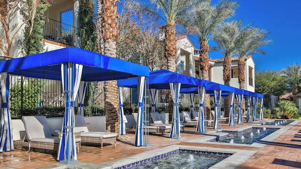 Un espace aménagé au bord de la piscine, avec des chaises longues et des parasols, entouré de palmiers et d’un bâtiment doté d’un toit en tuiles.