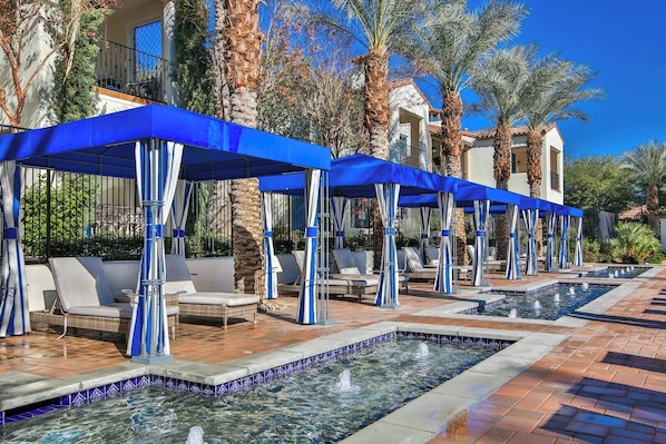 Ein Poolbereich mit Liegestühlen und Sonnenschirmen, umgeben von Palmen und einem Gebäude mit einem Ziegeldach.