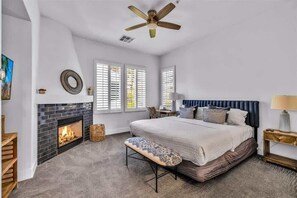 A bedroom with a fireplace, a bed with a blue headboard, a ceiling fan, and a window with blinds. - The Luxe Loft C | Studio Legacy Villa (La Quinta)