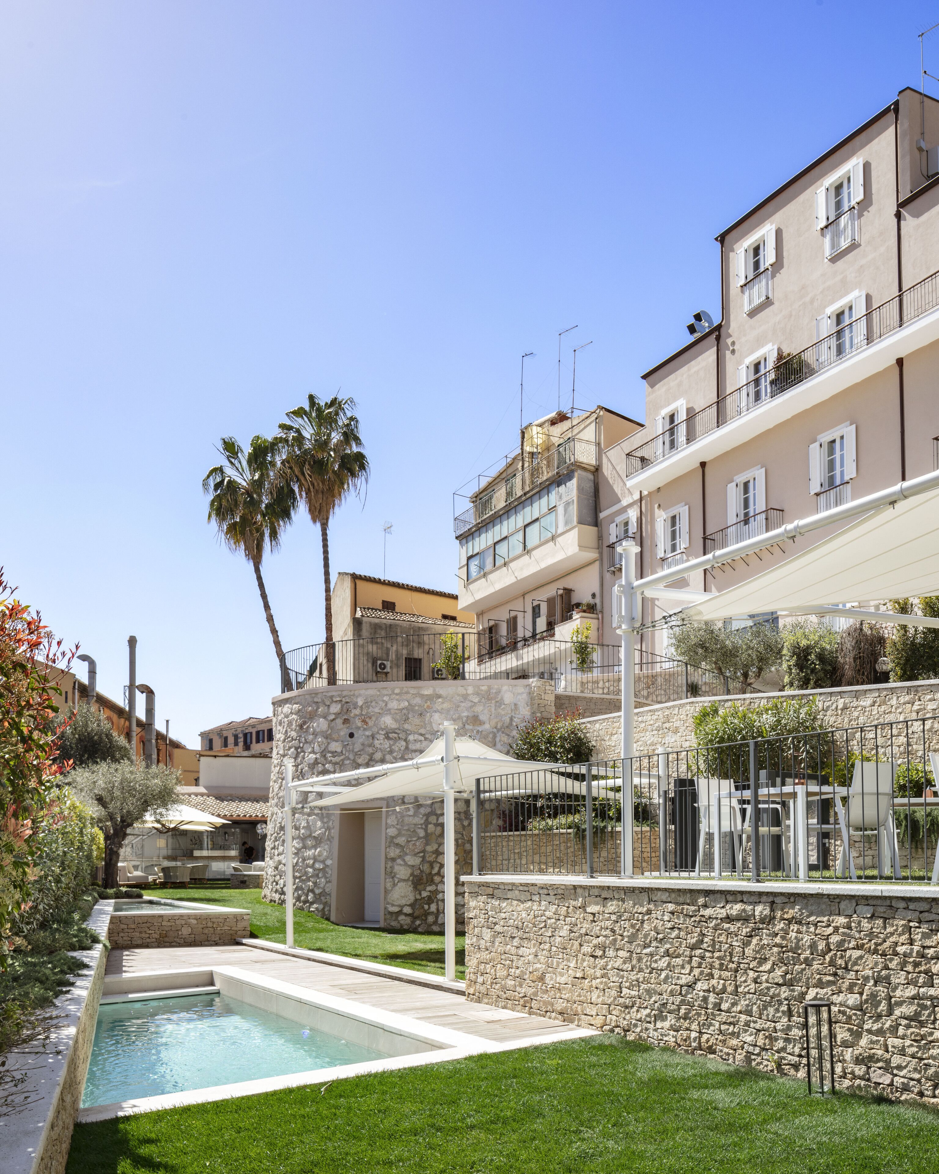 A residential building with a swimming pool, stone walls, and a garden with palm trees.